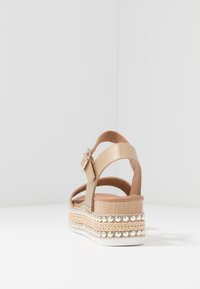 Beige Wedge-Sandal mit Knöchelriemen, Schnalle und dekorativen metallischen Nieten auf einer geschichteten Sohle vor weißem Hintergrund.