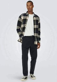 Camisa de flanela quadriculada em preto e bege, usada sobre uma t-shirt branca, combinada com calças escuras e tênis. Conjunto casual com design simples.