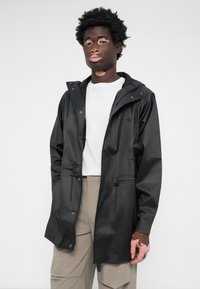 Veste noire imperméable avec capuche, cordons de serrage ajustables et deux poches zippées. Portée par-dessus une chemise blanche et un pantalon beige. Texture lisse.