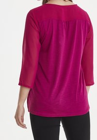 Top fucsia con corpo texturizzato e maniche trasparenti a 3/4. Presenta una scollatura rotonda e pieghe sulla parte posteriore della spalla. Abbinato a pantaloni neri.