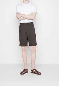 Brune shorts lavet af stof med knaplukning og to sidelommer, parret med mørkebrune snøresandaler. Enkel hvid T-shirt.