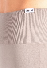 Beige geribbelde tailleband met een gladde textuur, voorzien van een wit label met "SMILODOX" aan de bovenrand van het kledingstuk.
