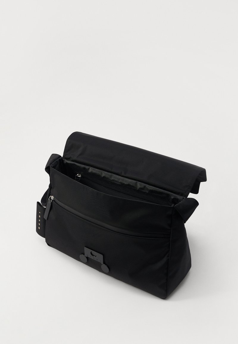 Sac en bandoulière en tissu noir avec fermeture à rabat, poche zippée à l'avant, texture douce et petite étiquette attachée pour le détail.