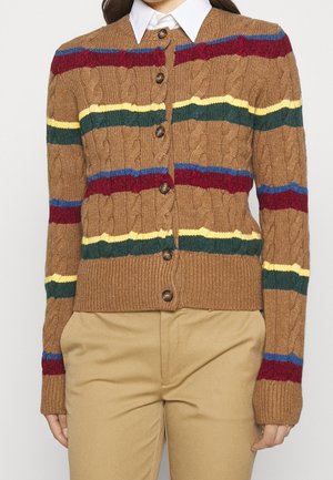 Cardigan marron en maille torsadée avec des rayures horizontales rouge, verte, jaune et bleue, fermeture boutonnée, porté sur une chemise blanche à col et un pantalon beige.