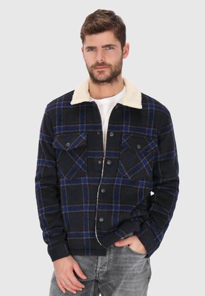 Mann trägt eine schwarz-blaue karierte Jacke mit weißem Sherpa-Kragen und grauen Jeans, steht mit einer Hand in der Tasche vor einem einfarbigen Hintergrund.