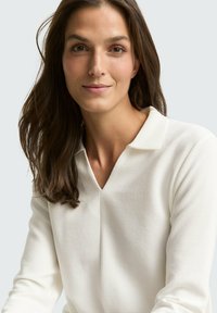 Weißes, geripptes langärmliges Oberteil mit einem V-Ausschnitt und einem Kragen, das eine glatte Textur aufweist. Das Model hat lange braune Haare.