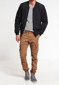 Czarna kurtka bomber, koszulka w paski, beżowe spodnie cargo, szare trampki. Bomber ma ściągacze przy mankietach, a spodnie cargo posiadają kieszenie boczne.