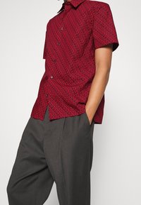 Camisa de manga corta en rojo con un patrón abstracto negro, que presenta un diseño de botones y un collar clásico, combinada con pantalones grises.