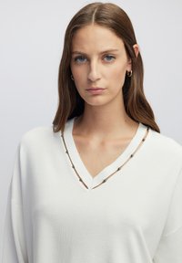 Witte gebreide trui met een V-hals, voorzien van een bruin accent langs de randen en versierd met zilveren kralen. Lange mouwen en een losse pasvorm. Soepele textuur.