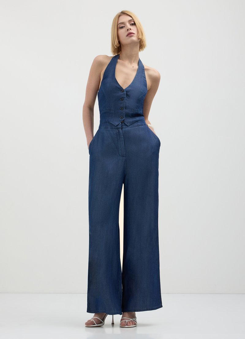La combinaison en denim présente un décolleté halter, une fermeture à boutons sur le devant et un design à jambes larges. Le tissu est d'un bleu profond avec une texture lisse.