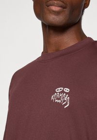 Sweatshirt em tons de bordeaux feito de um tecido macio, apresentando um logotipo gráfico branco no peito. Decote redondo com uma textura suave e um corte descontraído.