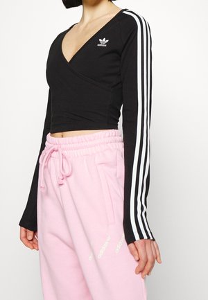 Kobieta ubrana w czarny, długi rękaw crop top Adidas z białymi paskami oraz różowe spodnie dresowe Adidas z wiązaniem w talii.