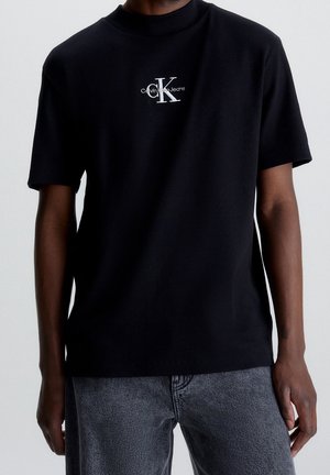 Černé tričko s krátkým rukávem s malým bílým logem CK Calvin Klein Jeans na hrudi, nošené se šedými džínami.