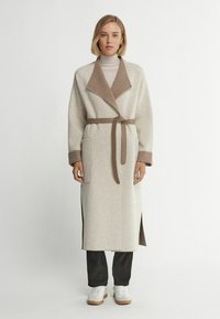 Beige inlindad kappa med en brun krage, sidoöppningar och bälte. Har två framfickor och en mjuk textur. Kombineras med mörka byxor och vita sneakers.