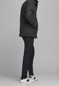 Cappotto di lana grigio scuro con colletto a bottoni, abbinato a pantaloni blu navy e sneaker bianche con dettagli blu e rossi.