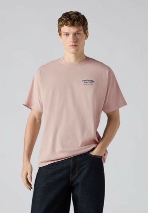 Joven con cabello rizado que lleva una camiseta rosa claro "Two Horse Aquatic Lounge" y vaqueros oscuros, con una mano en el bolsillo, fondo neutro.