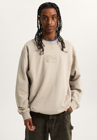 Beige cotton sweatshirt with embroidered Vans logo, round neck, dropped shoulders, and ribbed cuffs. Worn with dark patterned pants.