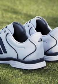 Ζευγάρι λευκά αθλητικά παπούτσια Adidas με μαύρες ρίγες τοποθετημένα σε πράσινο γρασίδι, σε θέα από πίσω και από το πλάι.