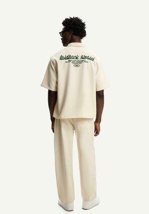 Beige short-sleeve shirt and matching pants, featuring green embroidered text on the back, standing figure with glasses and curly hair.