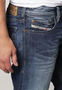 Jeans de denim azul oscuro con un acabado desgastado. Las características incluyen un patrón de costura visible, múltiples bolsillos y una etiqueta de marca en la cinturilla.