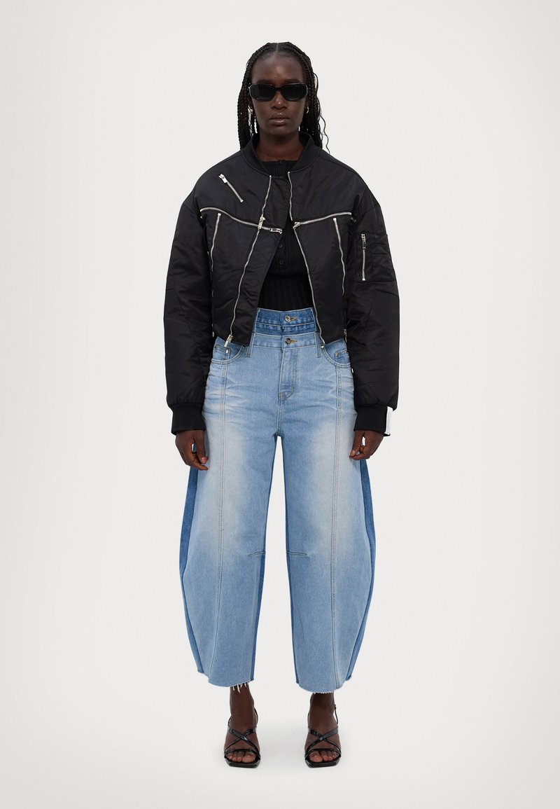Zwarte cropped bomberjack met zilveren ritsen, gecombineerd met lichtblauwe high-waisted baggy jeans met een denimpaneel. Transparante zonnebril.