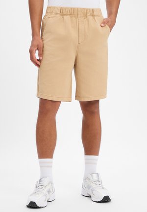 Uomo che indossa pantaloncini beige con vita elastica, mani nelle tasche, calzini bianchi e scarpe da corsa bianche, in piedi su uno sfondo semplice.
