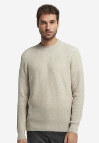 Beige gerippter Strickpullover mit Rundhalsausschnitt und langen Ärmeln, der ein strukturiertes Finish und eine lockere Passform aufweist, kombiniert mit dunklen Jeans.