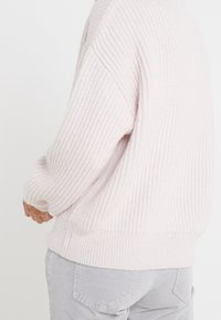 Różowy sweter o prążkowanej strukturze, luźny krój i opadające ramiona, z miękką teksturą i szerokim paskiem w talii, w zestawie z jasnoszarymi spodniami.