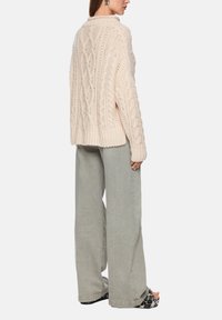 Grober Strickpullover in hellem Beige mit strukturierten Zopfmustern, lockerer Passform, fallenden Schultern und gerippten Bündchen, kombiniert mit weiten grauen Hosen.