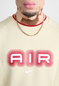 Sudadera beige con ribetes rojos que presenta el texto "AIR" en una fuente estilizada roja. Un collar de cadena de plata y oro acentúa el escote.