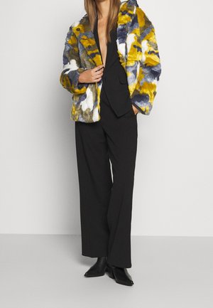 Vrouw draagt een meerkleurige nepbontjas over een zwart pak met wijde broekspijpen en zwarte laarzen met hakken, staand tegen een effen achtergrond.