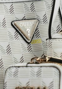 Sac à main blanc à motif, présentant un design en chevrons, un logo métallique triangulaire en argent et des accents dorés sur la tirette de la fermeture éclair.