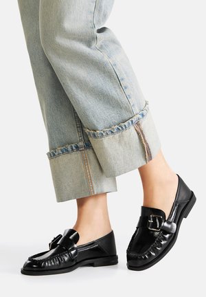 Schwarze Lederloafers mit strukturiertem Finish, ausgestattet mit einer dekorativen Schnalle und Stickdetails, kombiniert mit hochgekrempelten hellblauen Jeans.