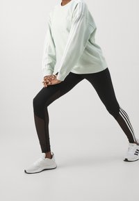 Light green sweatshirt with white three stripes, paired with black leggings featuring sheer panels and white athletic shoes.