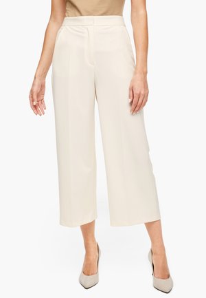 Mujer con pantalones crema de cintura alta y pierna ancha, recortados por encima de los tobillos, combinados con tacones puntiagudos a juego.
