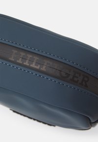 Borsa in materiale sintetico blu, liscio, con una striscia nera di accento che presenta la scritta in rilievo "HILFIGER" e dettagli di cucitura a contrasto.