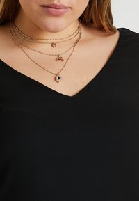 Collares en tonos dorados de capas presentan varios colgantes: un diseño en forma de ojo, un pequeño cangrejo y un charm circular. Llevados sobre una blusa negra.