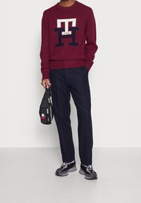 Pull en maille bordeaux avec logo blanc et bleu marine, associé à un pantalon bleu marine. Sac banane en tissu noir et baskets grises avec accents noirs.