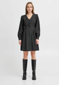 Robe boutonée gris foncé avec un décolleté en V, des manches longues bouffantes et un corsage ajusté, tombant au-dessus du genou. Associée avec des bottes noires montantes.