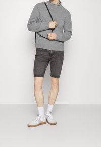 En person står i en grå tröja och mörka shorts, håller i en svart rem, bär vita strumpor och sneakers, mot en ljus bakgrund.