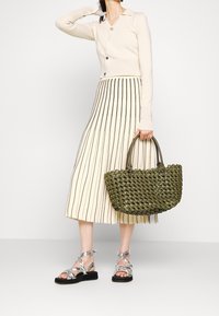 Bolso de mano verde tejido con dos asas, cárdigan crema con botones, falda midi de rayas en beige y negro, y sandalias plateadas con tiras.