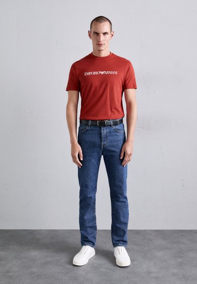 Tricou roșu din bumbac cu logo-ul alb "Emporio Armani", asortat cu blugi albaștri și adidași albi. Modelul stă drept cu mâinile relaxate.