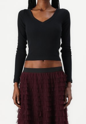 Frau trägt ein schwarzes, langärmeliges, bauchfreies V-Ausschnitt-Oberteil und einen mehrlagigen, bordeauxroten Rock mit Rüschen, gezeigt von Hals bis Mitte Oberschenkel.