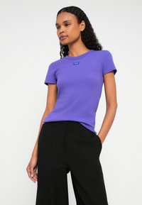 Donna con capelli ricci che indossa una maglietta aderente viola a maniche corte e pantaloni neri larghi, in piedi contro uno sfondo bianco semplice.