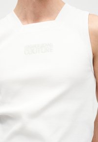 Gros plan d'une personne portant un haut blanc sans manches avec le logo en relief "Versace Jeans Couture" sur la poitrine.