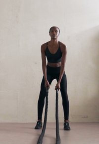 Žena u crnoj sportskoj odjeći vježba s bojama užadima u minimalnom unutarnjem prostoru s jednostavnom svijetlom pozadinom zida.