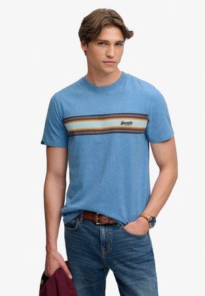 Ung mand iført en blåstribet T-shirt og jeans, der holder en foldet vinrød jakke i den ene hånd og den anden hånd i jeanslommen.