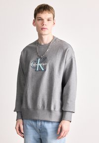 Šedá mikina s žebrovanými manžetami a lemem, s logem "Calvin Klein" v modré a bílé. Model nosí s světle modrými džíny a stříbrným řetězem.