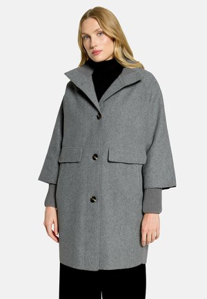 Femme blonde aux yeux bleu clair portant un manteau en laine gris avec de gros boutons par-dessus un pull à col roulé noir.