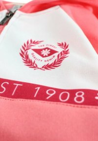 Chemise à manches courtes rouge et blanche arborant un logo avec des feuilles de laurier, "THE FINEST QUALITY DENIM" et "EST 1908" imprimés en rose.